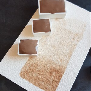 K&ouml;nnte beinhalten: Drei braune Aquarellfarben in wei&szlig;en Kunststoffbeh&auml;ltern sind auf wei&szlig;em Aquarellpapier mit einem braunen Farbfleck angeordnet. Die Farbe hat einen satten, erdigen Ton, und das Papier hat eine strukturierte Oberfl&auml;che.
