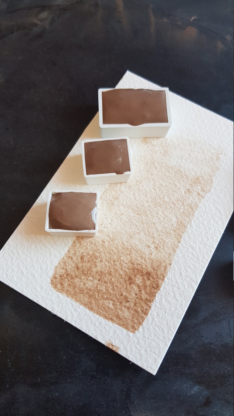 K&ouml;nnte beinhalten: Drei braune Aquarellfarben in wei&szlig;en Kunststoffbeh&auml;ltern sind auf wei&szlig;em Aquarellpapier mit einem braunen Farbfleck angeordnet. Die Farbe hat einen satten, erdigen Ton, und das Papier hat eine strukturierte Oberfl&auml;che.