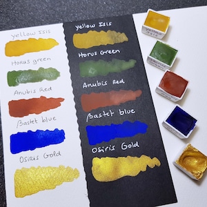 Pode incluir: Uma cole&ccedil;&atilde;o de tintas aquarela em v&aacute;rias cores, incluindo amarelo, verde, vermelho, azul e dourado. As cores s&atilde;o amostradas em papel branco e preto, com os nomes das cores escritos ao lado de cada amostra. Pequenos recipientes de tinta tamb&eacute;m s&atilde;o vis&iacute;veis.