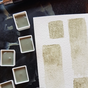 K&ouml;nnte beinhalten: Aquarellfarben in kleinen wei&szlig;en Beh&auml;ltern, neben Farbfeldern auf wei&szlig;em Papier. Die Farbe ist ein ged&auml;mpftes Gr&uuml;n mit sichtbarer Textur. Der Hintergrund ist eine dunkle Oberfl&auml;che.