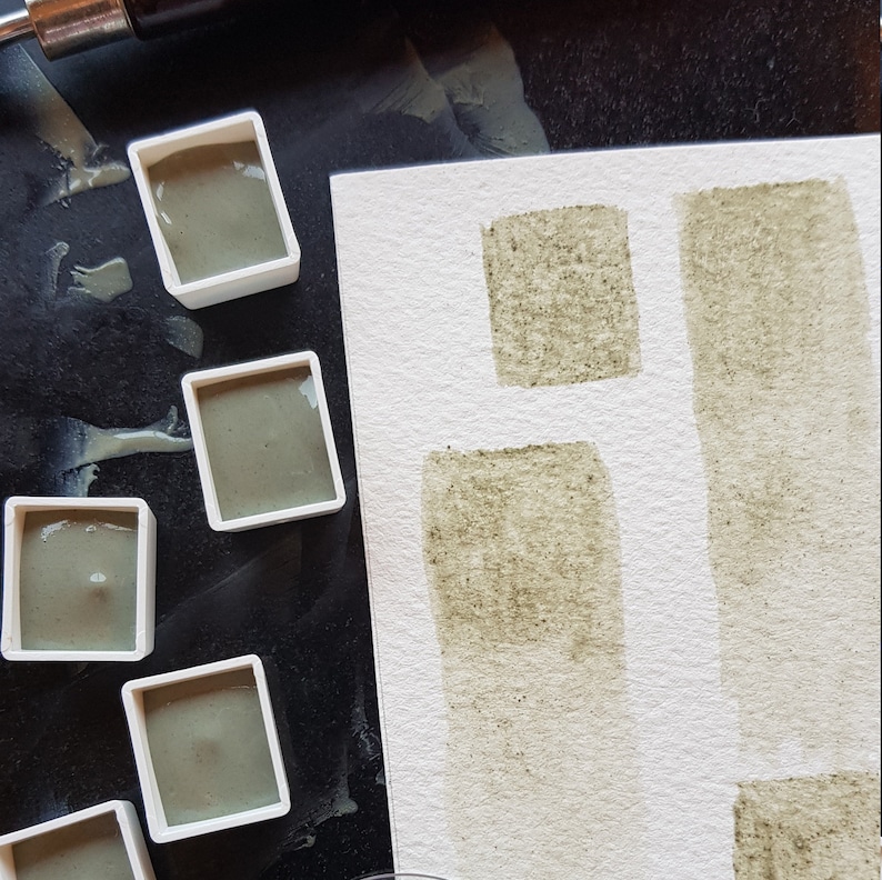 K&ouml;nnte beinhalten: Aquarellfarben in kleinen wei&szlig;en Beh&auml;ltern, neben Farbfeldern auf wei&szlig;em Papier. Die Farbe ist ein ged&auml;mpftes Gr&uuml;n mit sichtbarer Textur. Der Hintergrund ist eine dunkle Oberfl&auml;che.