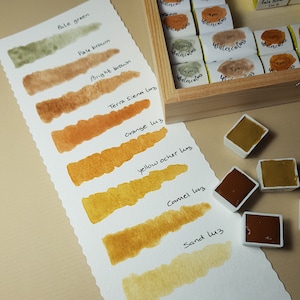 K&ouml;nnte beinhalten: Ein Aquarellfarbenset mit einer Holzbox und einer Farbkarte. Die Karte zeigt Farbfelder in Erdt&ouml;nen, darunter blasse Gr&uuml;n-, Braun- und Oranget&ouml;ne. Einzelne Aquarelln&auml;pfchen sind ebenfalls sichtbar, die das Pigment der Farbe zeigen.