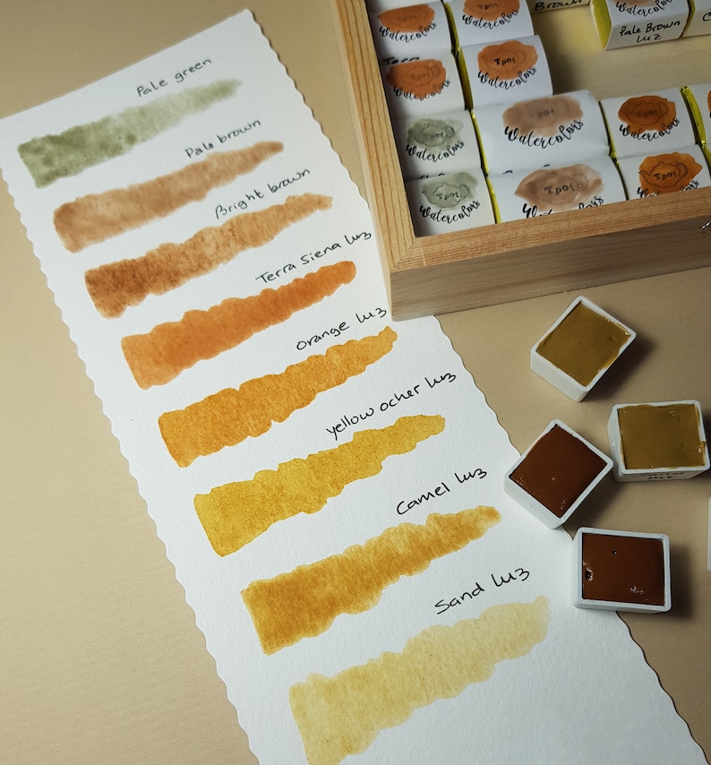 K&ouml;nnte beinhalten: Ein Aquarellfarbenset mit einer Holzbox und einer Farbkarte. Die Karte zeigt Farbfelder in Erdt&ouml;nen, darunter blasse Gr&uuml;n-, Braun- und Oranget&ouml;ne. Einzelne Aquarelln&auml;pfchen sind ebenfalls sichtbar, die das Pigment der Farbe zeigen.