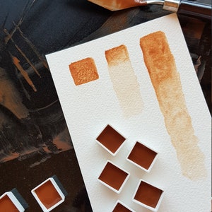 K&ouml;nnte beinhalten: Draufsicht auf Aquarellfarben und einen Malmesser. Die Farben sind in einem satten Braunton gehalten und werden in kleinen quadratischen Beh&auml;ltern und auf Papier pr&auml;sentiert. Ein Malmesser mit brauner Farbe liegt auf einer dunklen Oberfl&auml;che.