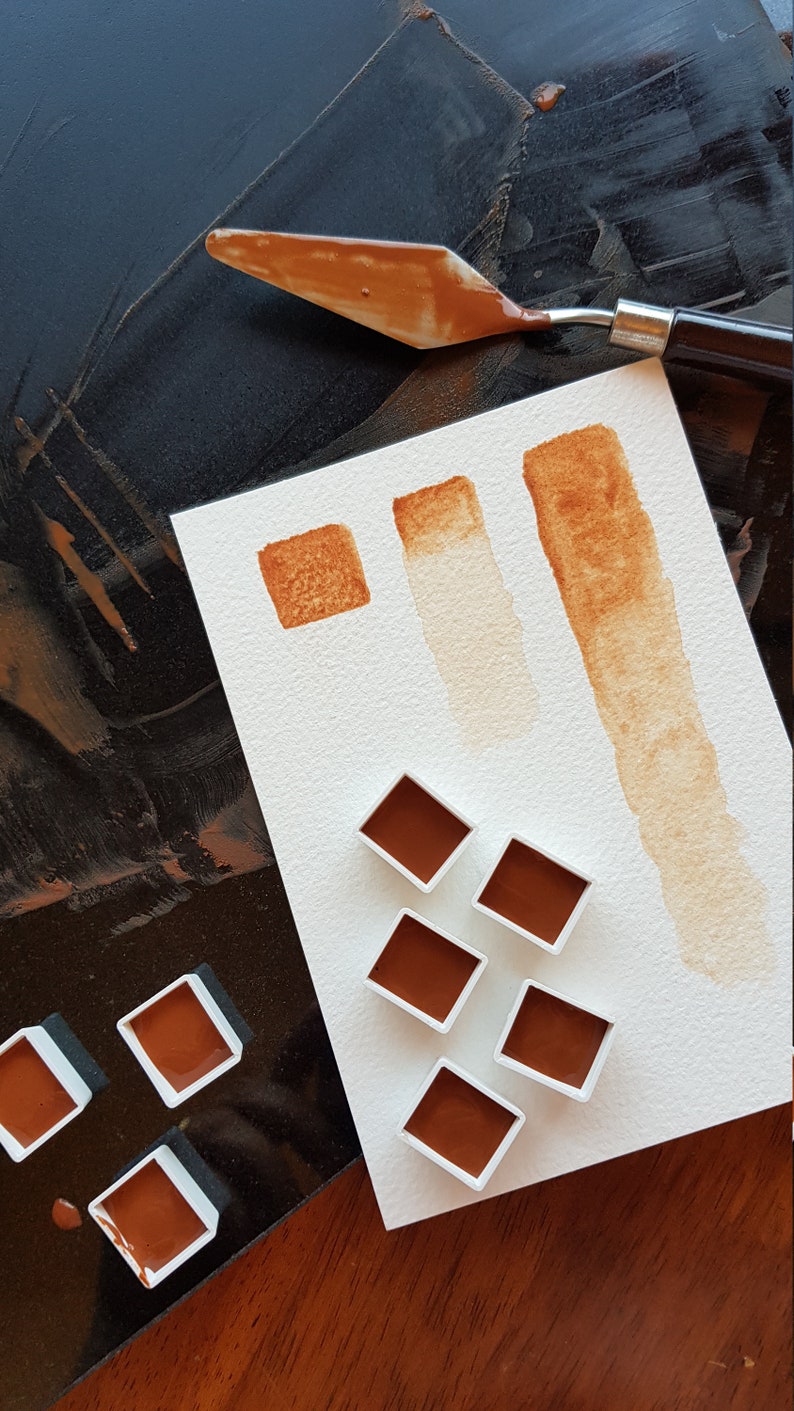 K&ouml;nnte beinhalten: Draufsicht auf Aquarellfarben und einen Malmesser. Die Farben sind in einem satten Braunton gehalten und werden in kleinen quadratischen Beh&auml;ltern und auf Papier pr&auml;sentiert. Ein Malmesser mit brauner Farbe liegt auf einer dunklen Oberfl&auml;che.