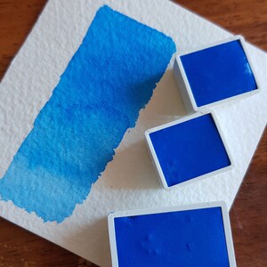 May include: A close-up of watercolor paints and a swatch. Three square, cobalt blue watercolor paint pans sit on a white surface next to a blue watercolor paint swatch. The swatch shows a gradient of blue hues.
