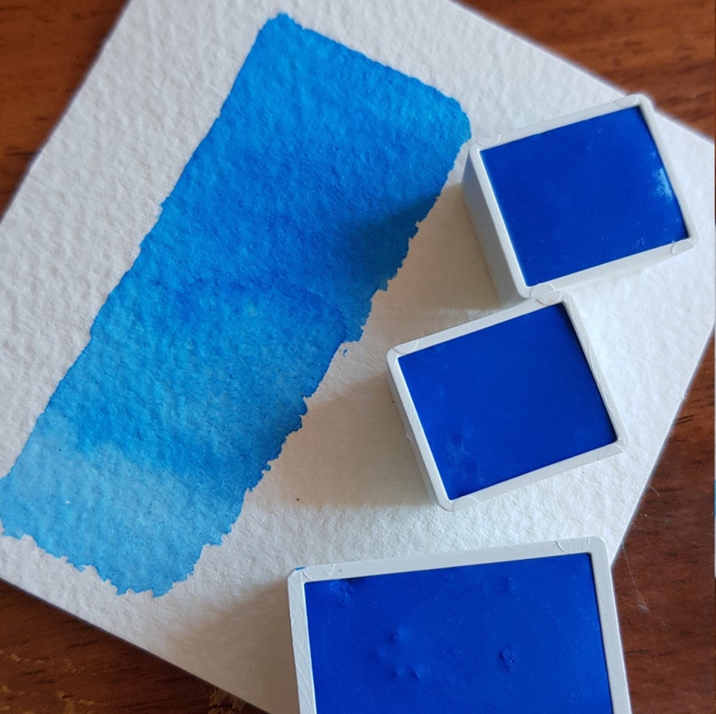 May include: A close-up of watercolor paints and a swatch. Three square, cobalt blue watercolor paint pans sit on a white surface next to a blue watercolor paint swatch. The swatch shows a gradient of blue hues.