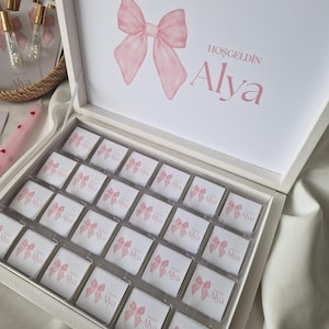 Può includere: Una scatola regalo bianca piena di cioccolatini confezionati singolarmente. Ogni involucro di cioccolato presenta un fiocco rosa e il nome "Alya". Il coperchio della scatola mostra un grande fiocco rosa e il testo "HOŞGELDİN Alya".
