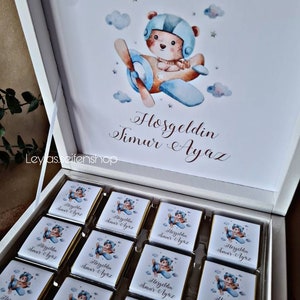 Peut inclure: Une boîte blanche avec un couvercle ouvert, révélant un plateau de petites barres de chocolat. Chaque barre a une image d'un ours en dessin animé portant un casque de pilote et pilotant un avion. Le texte "Hoşgeldin Timur Ayaz" est imprimé sur le couvercle de la boîte.