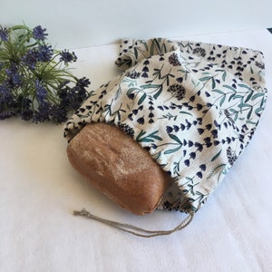 Könnte beinhalten: Eine weiße Leinentasche für Brot mit einem Blumenmuster in Grün- und Lilatönen. Die Tasche ist oben zusammengezogen und mit einer Schnur gebunden. Ein Brotlaib befindet sich in der Tasche.