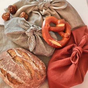 Könnte beinhalten: Nahaufnahme von drei in Stoff verpackten Paketen. Die Pakete bestehen aus Naturleinen und einem dunkelbraunen Stoff. Die Pakete enthalten ein Brot, ein Brezel und ein paar kleine Brötchen.