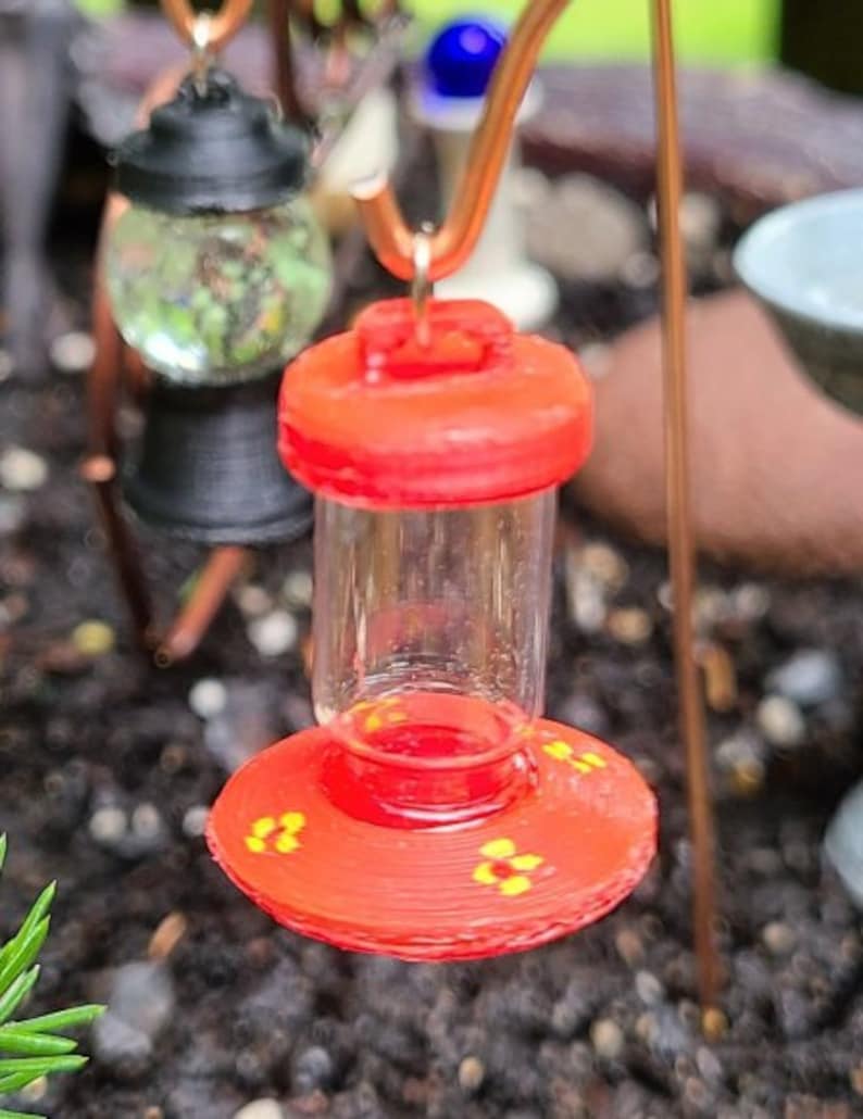 Pu&ograve; includere: Un alimentatore per colibr&igrave; rosso con un serbatoio di vetro trasparente e una base decorativa con accenti di fiori gialli.
