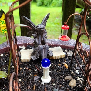 Pu&ograve; includere: Una scena di giardino delle fate in miniatura con una piccola statua di fata grigia seduta su una panchina, una casetta per uccelli gialla, un alimentatore per colibr&igrave; rosso e un lampione blu. Il giardino &egrave; in un vaso marrone con terra e pietre.