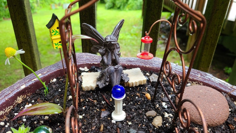Pu&ograve; includere: Una scena di giardino delle fate in miniatura con una piccola statua di fata grigia seduta su una panchina, una casetta per uccelli gialla, un alimentatore per colibr&igrave; rosso e un lampione blu. Il giardino &egrave; in un vaso marrone con terra e pietre.