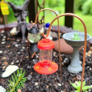 Pu&ograve; includere: Un mini-alimentatore rosso per colibr&igrave; con un contenitore di vetro trasparente e accenti di fiori gialli &egrave; appeso a un supporto in filo di rame in un giardino.