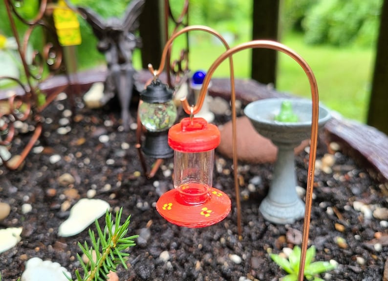 Pu&ograve; includere: Un mini-alimentatore rosso per colibr&igrave; con un contenitore di vetro trasparente e accenti di fiori gialli &egrave; appeso a un supporto in filo di rame in un giardino.