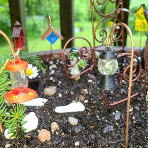 Pu&ograve; includere: Una scena da giardino in miniatura con un piccolo albero sempreverde, un mangiatoia per uccelli rossa, una lanterna nera con un globo di vetro trasparente e un abbeveratoio per uccelli in ceramica bianca con una statuetta di rana verde.