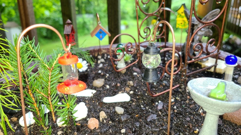 Pu&ograve; includere: Una scena da giardino in miniatura con un piccolo albero sempreverde, un mangiatoia per uccelli rossa, una lanterna nera con un globo di vetro trasparente e un abbeveratoio per uccelli in ceramica bianca con una statuetta di rana verde.