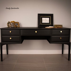 May include: A black wooden desk with six drawers and gold knobs. The desk has a simple design with straight lines and a rectangular shape. The desk is sitting on a grey tiled floor. The text "Simply Spectacular" is written on the wall behind the desk.