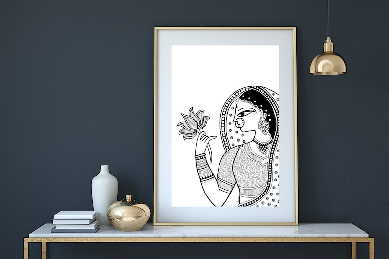 May include: Black and white line art illustration of a woman in traditional Indian clothing holding a lotus flower. The woman is wearing a headscarf, a necklace, and bangles. The artwork is framed and displayed on a white wall.