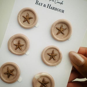 May include: Six beige wax seals with a bronze starfish design are arranged on a white card. The seals are round with a raised starfish in the centre. The card has the text "Handmade in Canada" and "BAY & HARBOUR" in black script.