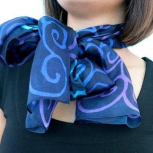 May include: A dark blue silk scarf featuring a swirling, abstract pattern in shades of blue and purple. The scarf is knotted around the neck, displaying its vibrant colours and design. Scarf dimensions are not visible in the image.