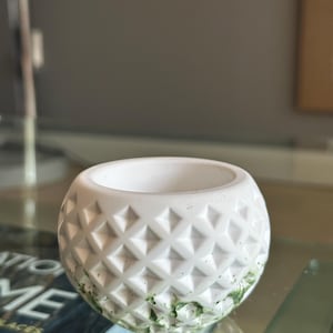 May include: A small, white, round planter with a geometric diamond pattern. The planter has a smooth interior and a textured exterior. Green moss is visible near the base. Suitable for small succulents or other decorative plants.