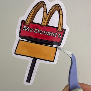 May include: A sticker of the McDonald's logo, featuring the golden arches and the name "McDonald's" in red lettering. The sticker also includes a yellow sign. The sticker is being held by a pair of tweezers.