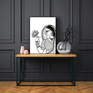 May include: Black and white line art print of a woman in traditional Indian clothing holding a lotus flower. The woman is wearing a veil and jewelry. The print is framed and displayed on a wooden table.