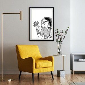May include: Black and white line art print of a woman holding a lotus flower. The print is framed in a black frame and hanging on a white wall.