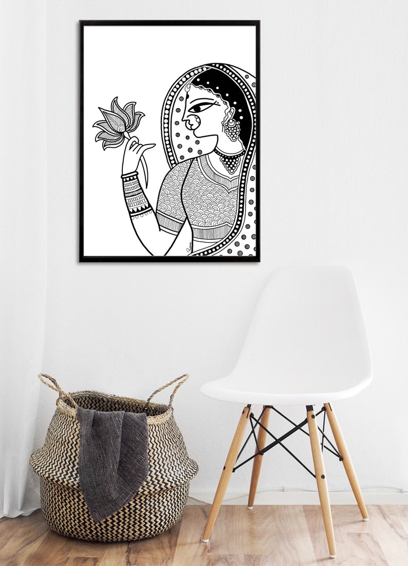 May include: Black and white line art print of a woman in traditional Indian clothing holding a lotus flower. The woman is wearing a patterned sari and jewelry. The print is framed in a black frame.