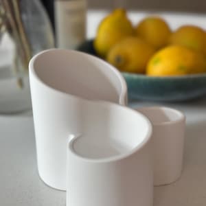 Puede incluir: Un soporte blanco de cerámica para utensilios con un diseño curvo único. El soporte tiene dos compartimentos y un acabado liso y mate. En el fondo, un cuenco de limones amarillos.