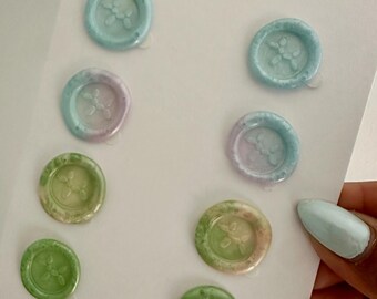 May include: A collection of colorful wax seals in shades of green, blue, and purple, each with a floral design. The seals are arranged on a white card with the text "Handmade in Canada" and "Bay & Harbour".