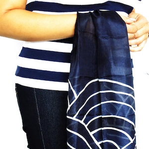 May include: A navy blue scarf with a white wave pattern is held in front of a person wearing a navy and white striped top and dark blue jeans. The scarf's design features curved white lines against a dark blue background.