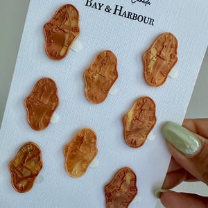 May include: Eight bronze-colored, melting clock-shaped pins on a white card. The pins are attached with small white adhesive squares. The card has the text "Handmade in Canada" and "BAY & HARBOUR" in black script.