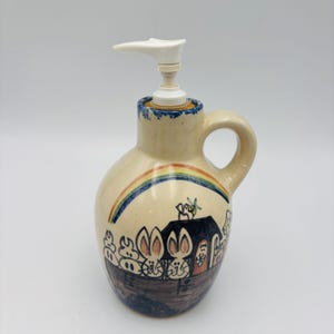May include: A ceramic soap dispenser with a white pump top and a handle. The beige dispenser features a hand-painted rainbow, a house, and cartoon rabbits. The rim is trimmed with blue paint.