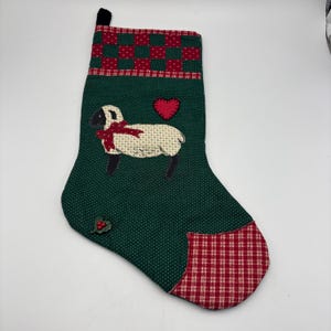 May include: A Christmas stocking with a green background and a sheep design. The stocking has a red and green checkered top and a red heart. The sheep has a red bow.