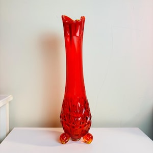 May include: A tall, red glass vase with a textured, diamond-shaped pattern. The vase has a flared opening and three short, rounded feet.