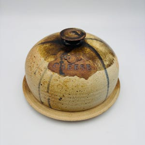 May include: A ceramic cheese dome and base. The dome is round with a small handle and a brown and tan colour scheme with dark brown accent lines. The word "CHEESE" is embossed on the front of the dome.