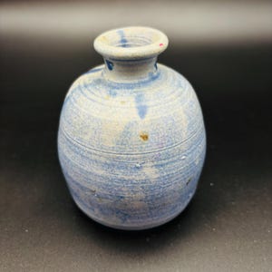 Puede incluir: Un pequeño jarrón de cerámica hecho a mano con cuerpo redondeado y cuello estrecho. Presenta un patrón arremolinado en azul claro y blanco, con sutiles motas marrones y azules. El borde es de un azul más claro.