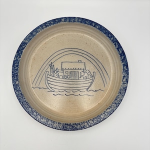 Puede incluir: Plato redondo de cerámica con centro beige y borde azul. El plato presenta una ilustración azul del Arca de Noé con animales y un arcoíris. El borde tiene un diseño moteado.