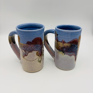 May include: Two handmade ceramic mugs with a tall, cylindrical shape. The mugs feature a blue rim and handle, with a blend of burgundy, tan, and lavender colors cascading down the sides. The mugs are designed for hot beverages.