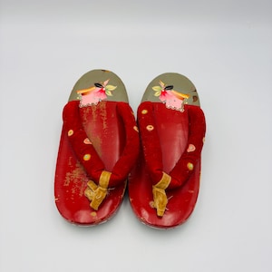 May include: Pair of traditional Japanese geta sandals. The wooden soles are painted red with a distressed finish. The straps are a deep red velvet with yellow accents and small decorative details. The top of the sandals are gray with floral designs.