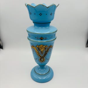 May include: A light blue, antique glass vase with a flared top and a round base. The vase features gold floral and scroll designs, with dark brown accents. The vase is approximately 10 inches tall.