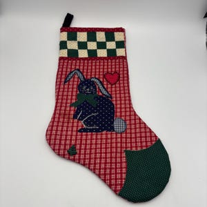 May include: A Christmas stocking with a red and white plaid pattern. It features an appliqued blue bunny with a green bow, a red heart, and a green and white checkered top. A small green tree is also appliqued on the stocking.