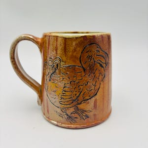 May include: A ceramic mug with a glossy, mottled amber and brown glaze featuring a hand-drawn black line illustration of a dodo bird. The mug has a curved handle on the left side and vertical, subtle darker brown stripes across the cylindrical body.
