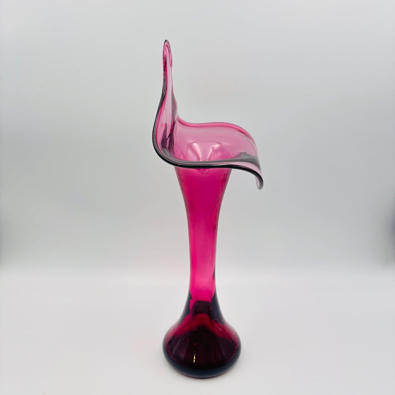 Puede incluir: Un jarr&oacute;n de vidrio rosa con un dise&ntilde;o curvo &uacute;nico. El jarr&oacute;n tiene una parte superior ancha y abierta y una base estrecha.