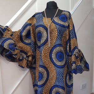 Könnte beinhalten: Ein langes, fließendes Kleid mit einem lebendigen blauen, braunen und weißen kreisförmigen Muster. Das Kleid hat breite, gerüschte Ärmel und ein passendes Muster. Eine silberne Halskette ist auf der Schaufensterpuppe drapiert.