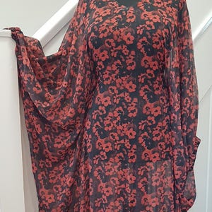 May include: A long-sleeved dress featuring a black base with a repeating red floral pattern, a V-neckline, and a ruffled hem. The garment is displayed on a mannequin and is paired with a chunky silver-toned chain link necklace at the neckline.