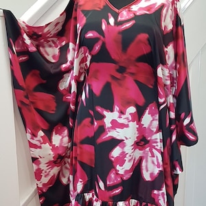 May include: A floor-length kaftan dress featuring a bold black, hot pink, and white floral pattern with a V-neckline and wide, flowing sleeves. The garment includes a gathered ruffle hem at the bottom and is displayed on a mannequin with a silver chain necklace.
